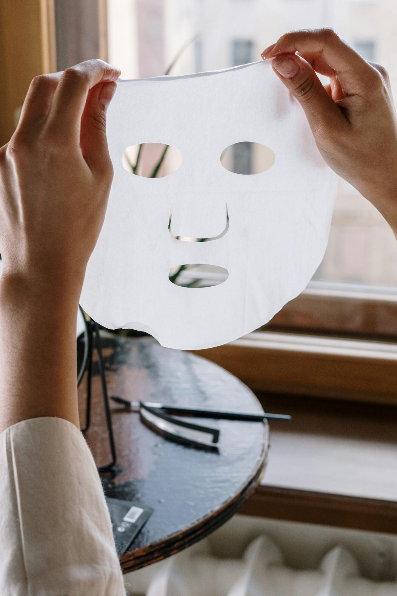 A woman holds a sheet mask in a cozy home setting, by a window.