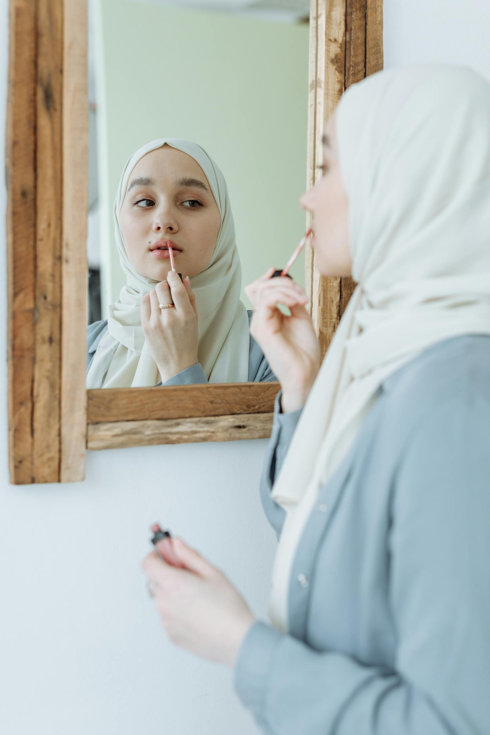 A woman in a hijab applying lipstick, captured in a natural setting, highlighting beauty and self-care.