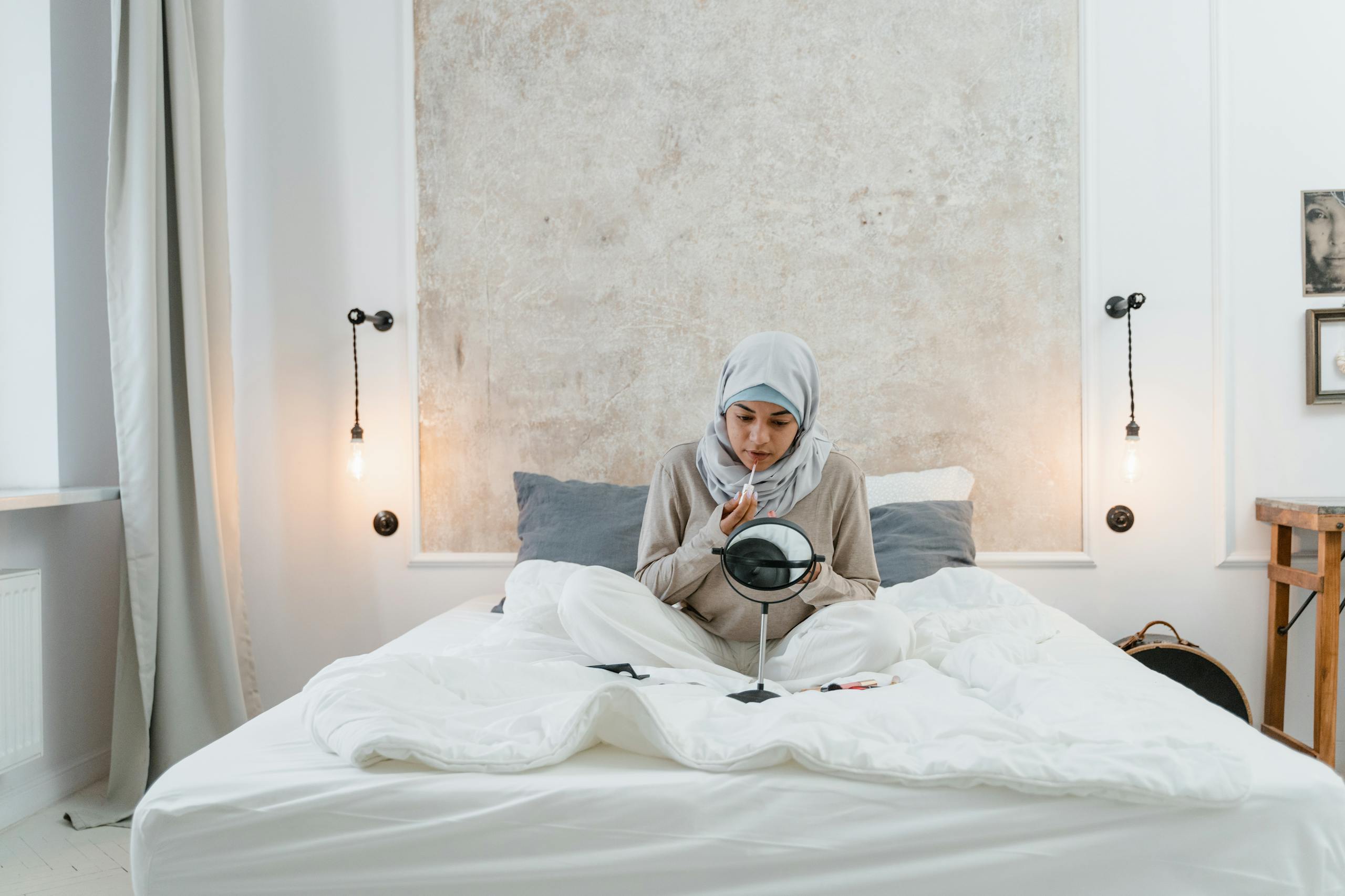 A woman wearing a hijab applies makeup while sitting on a bed in a cozy, well-lit bedroom.