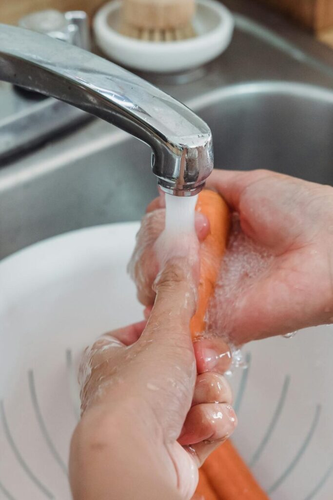 Person washing fresh carrots under a kitchen faucet using a colander, in a home setting.