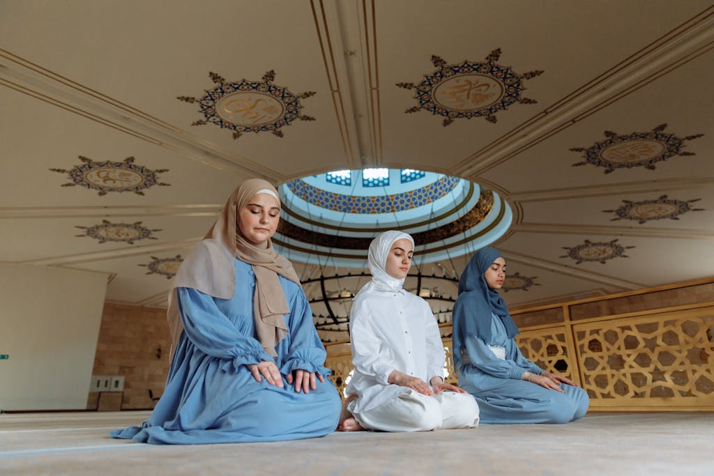 Three Muslim women in traditional attire praying inside a beautifully decorated mosque with intricate ceiling designs.