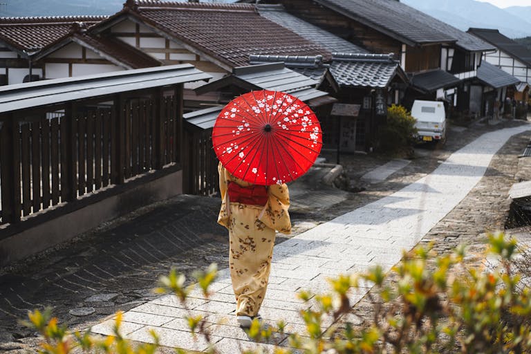 Woman in kimono with red umbrella walking through a traditional Japanese village street.