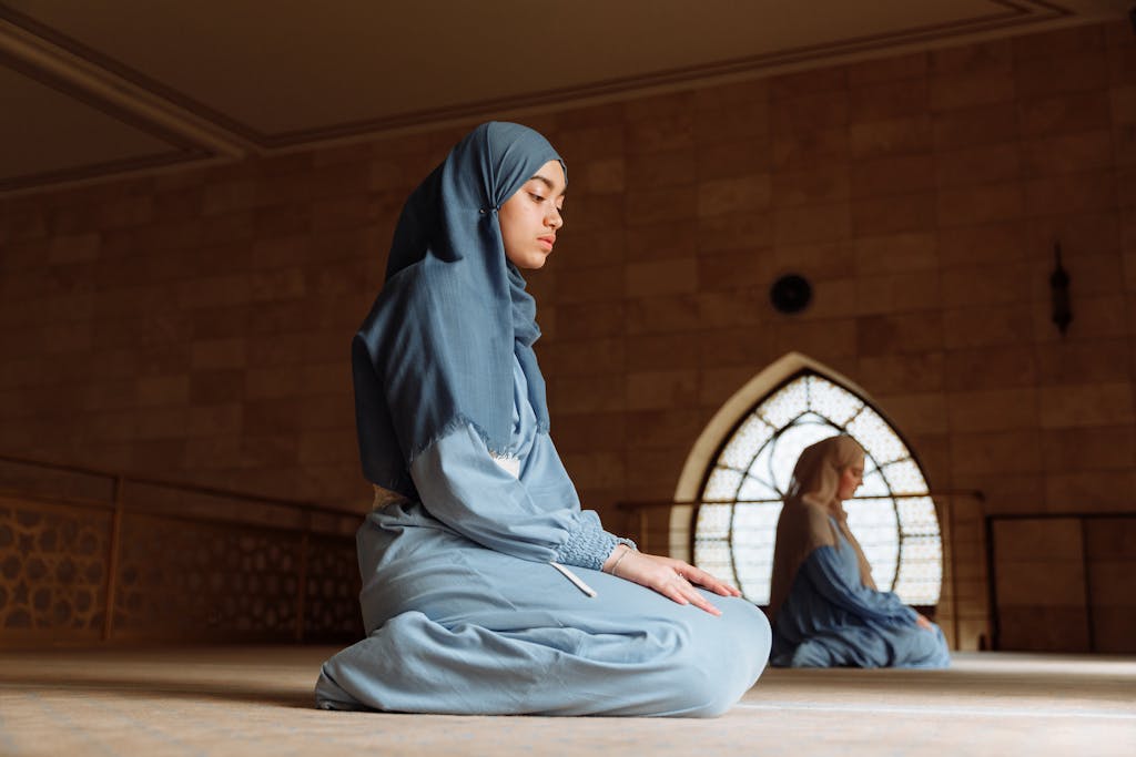 Women in hijab kneel in prayer inside a mosque, epitomizing devotion and tranquility.