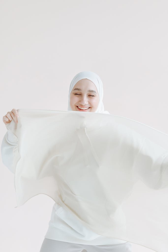 A smiling woman in a white hijab joyfully poses with flowing fabric in a minimalistic setting.