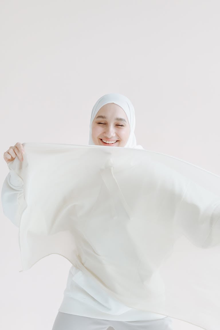 A smiling woman in a white hijab joyfully poses with flowing fabric in a minimalistic setting.