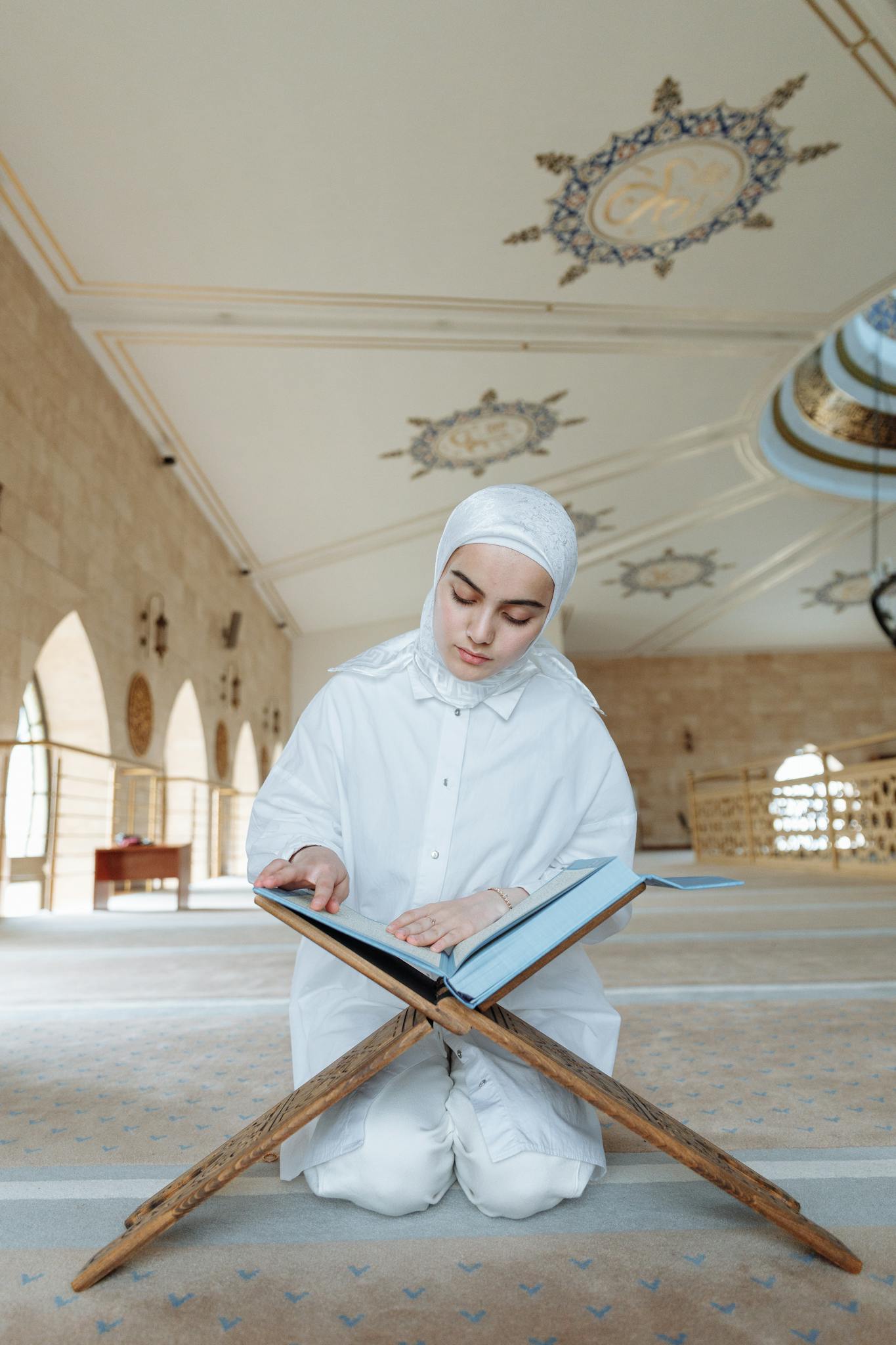 A woman in hijab kneels inside a mosque reading the Quran, capturing a moment of devotion.