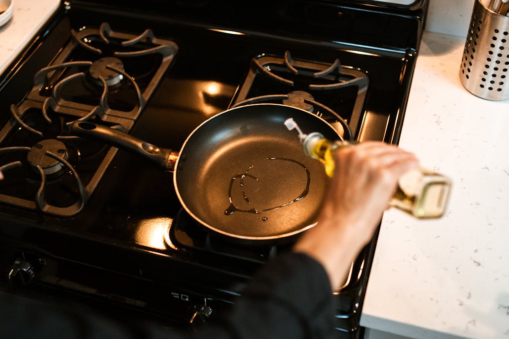 Close-up of person pouring olive oil into a frying pan on a kitchen stove.