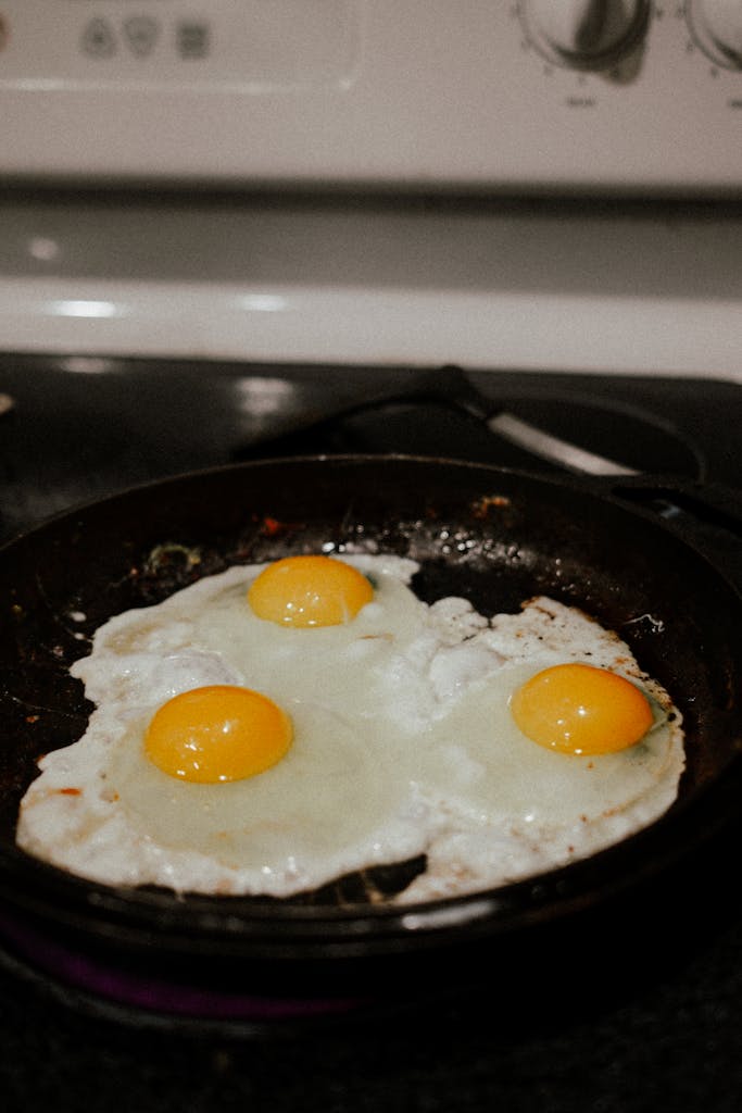 Close-up of three fried eggs sizzling in a skillet on a stovetop.