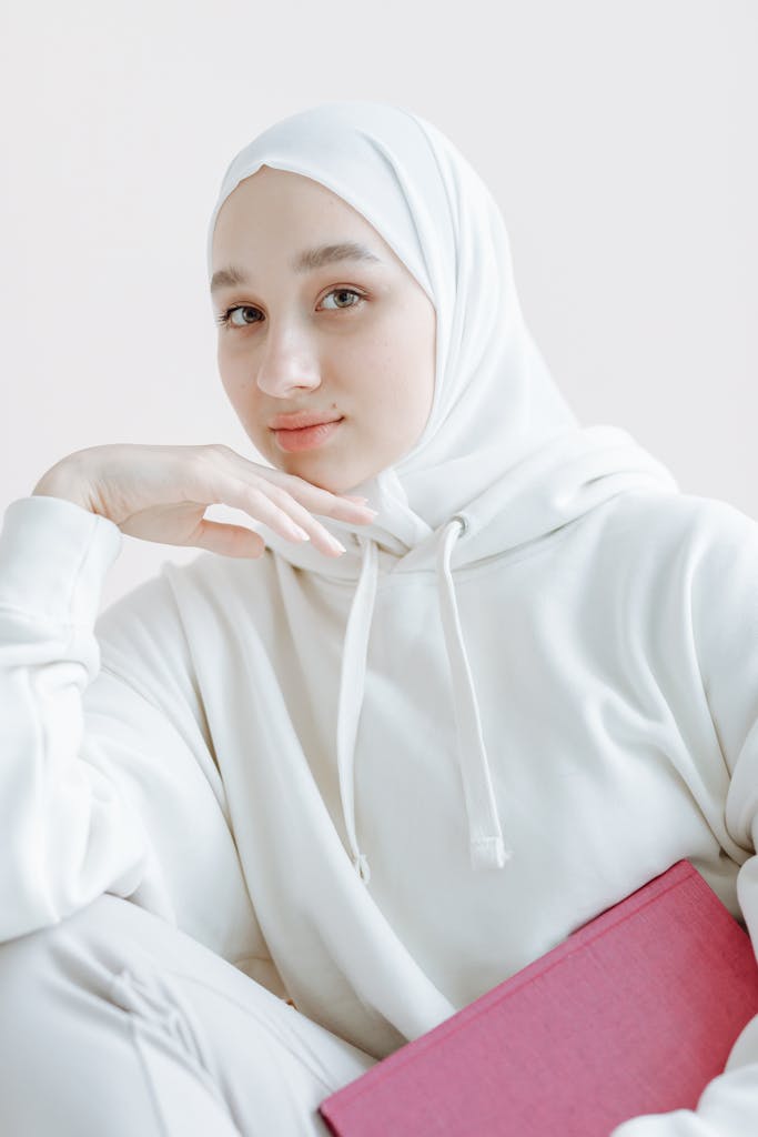 Portrait of a woman in a hijab and hoodie posing thoughtfully with a book, exuding calmness.