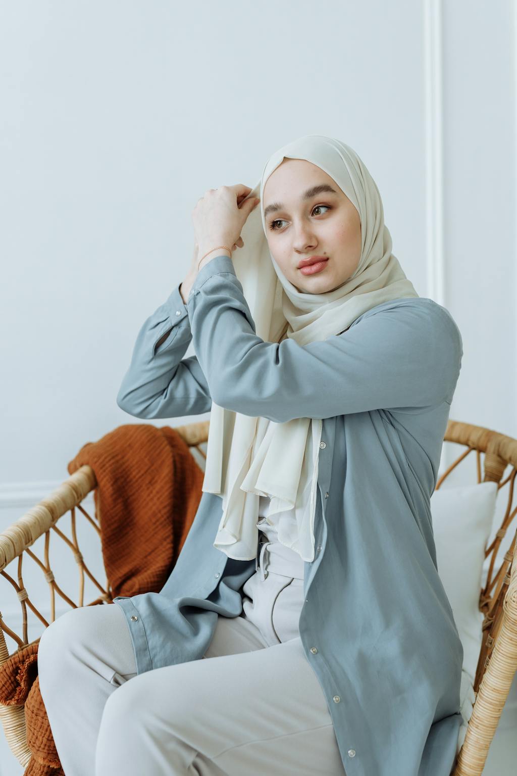 Portrait of a young Muslim woman adjusting her hijab while sitting indoors on a chair.