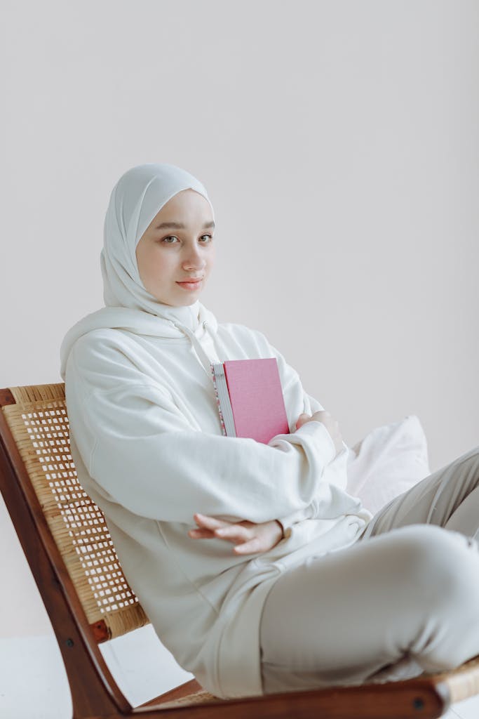 Woman in hijab sitting on rattan chair holding a notebook, embodying a peaceful lifestyle in a minimalist interior.